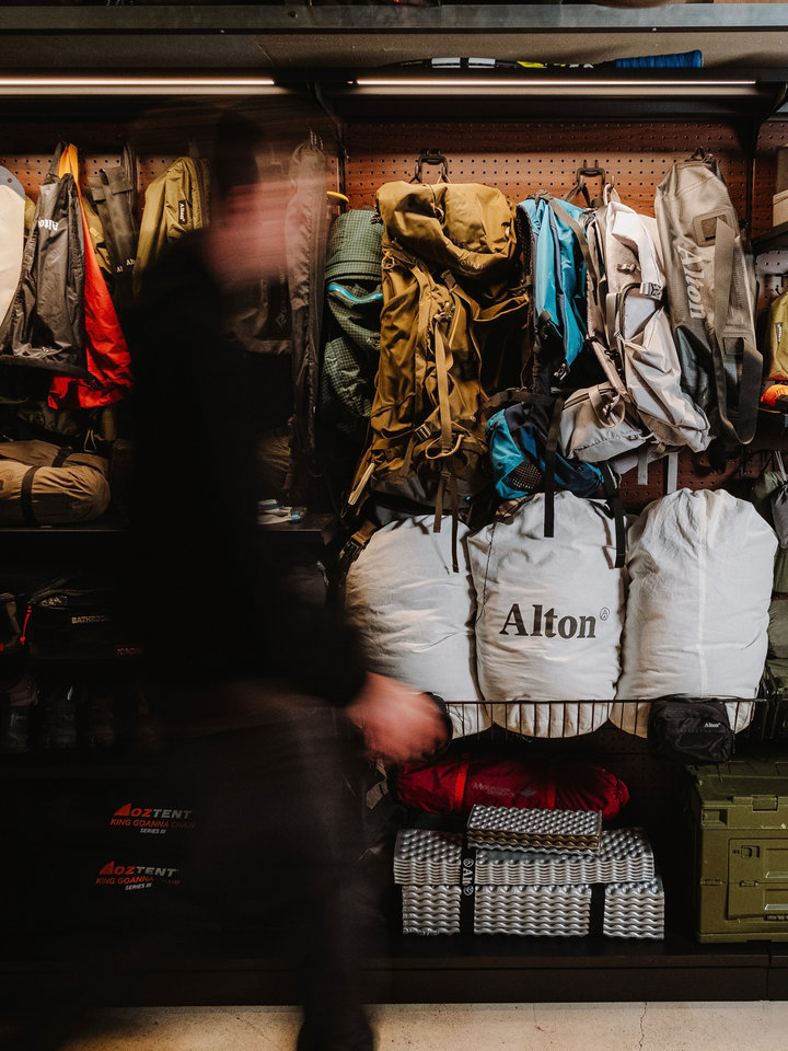 Backpacks, jackets and camping gear arranged on an illuminated PegGrid wall, with a blurred person passing in front.