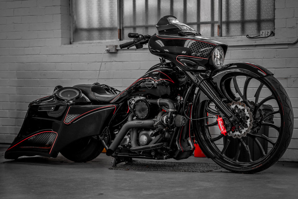 A monochrome custom touring motorcycle with red accents against a brick wall