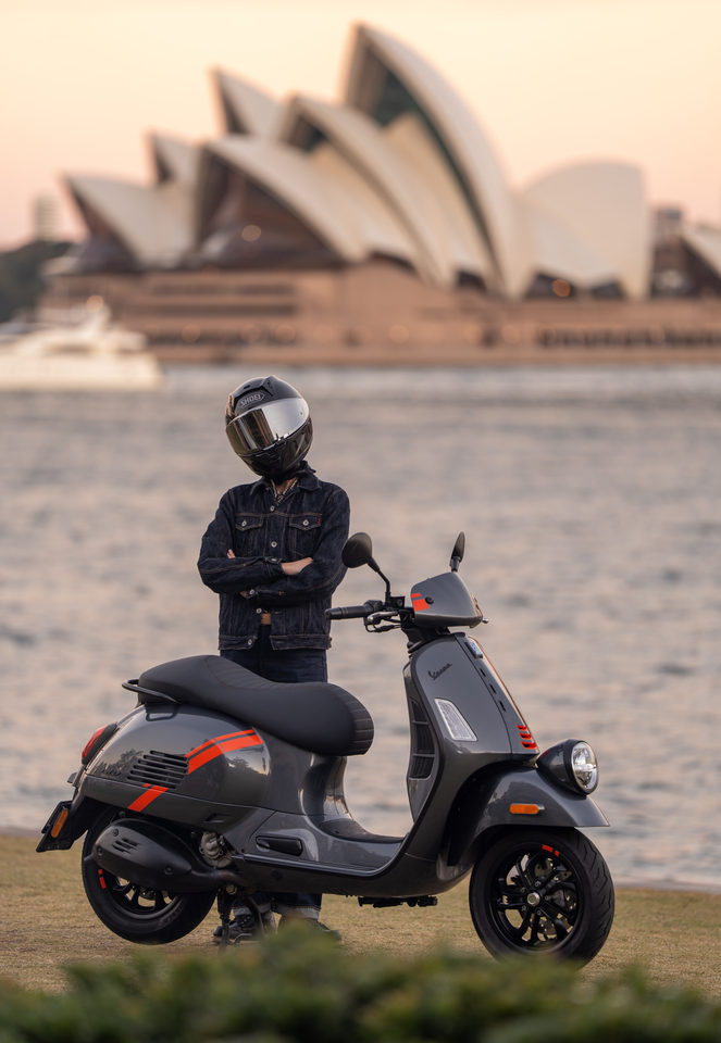 A helmeted rider beside a grey Vespa scooter with the Sydney Opera House across the water.